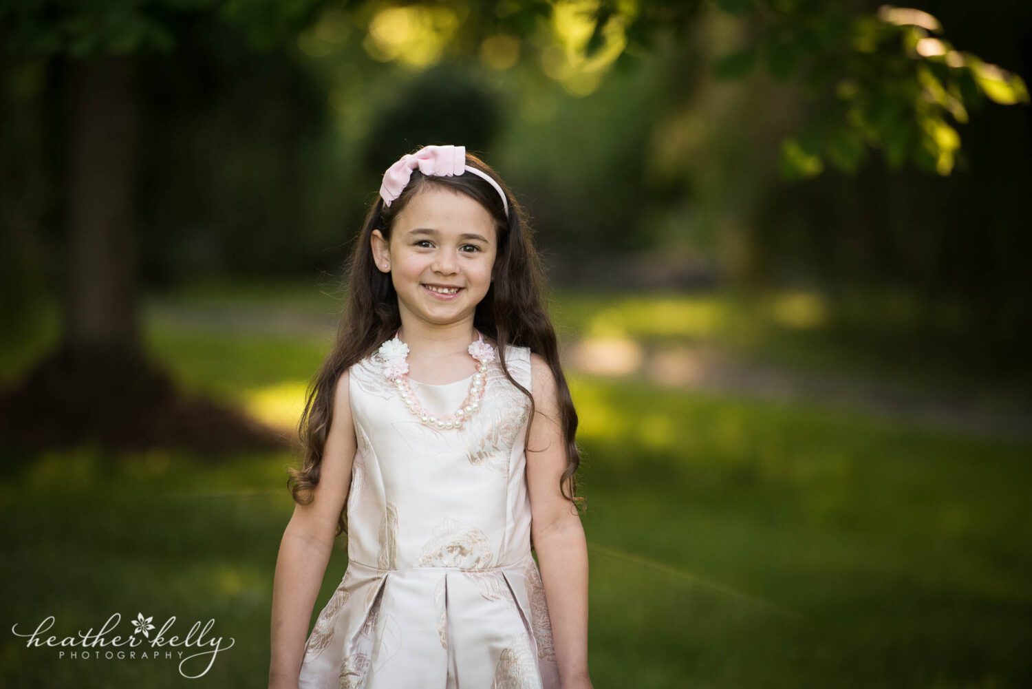 a girl in a pink dress and pink headband stands and smiles at the camera. Lush green trees are in the background for her monroe ct family photography session