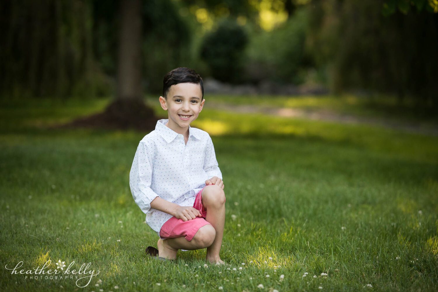 a boy in a white button down shirt and pink shorts kneels on the grass during his monroe ct family photography session