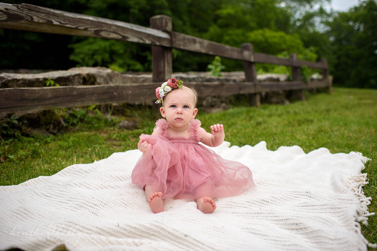 mahopac ny family photography

6 month old girl in pink dress and flower headband sits on a white blanket outside near a wooden fence. 