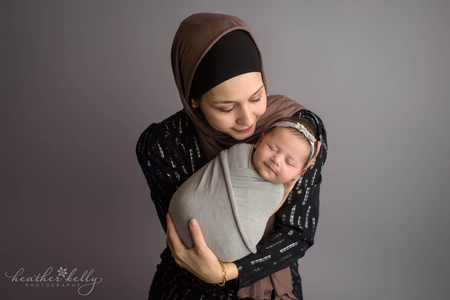 mom and newborn baby. Mom is holding a newborn girl that is wrapped in a gray swaddle. She is looking down at her in a newborn photography pose