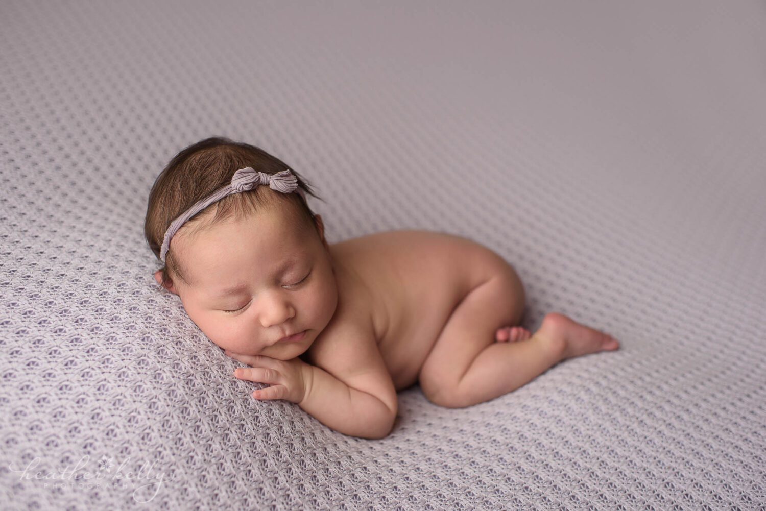 newborn photography of a sleeping newborn girl on a light purple backdrop with a purple bow in her hair