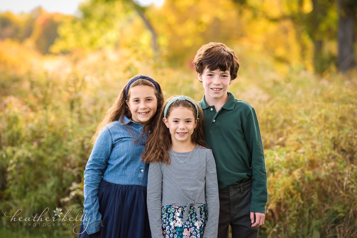 2022 fall mini sessions ct. Three siblings, two girls and older brother standing near each other looking at the camera. Pretty fall colors in the background