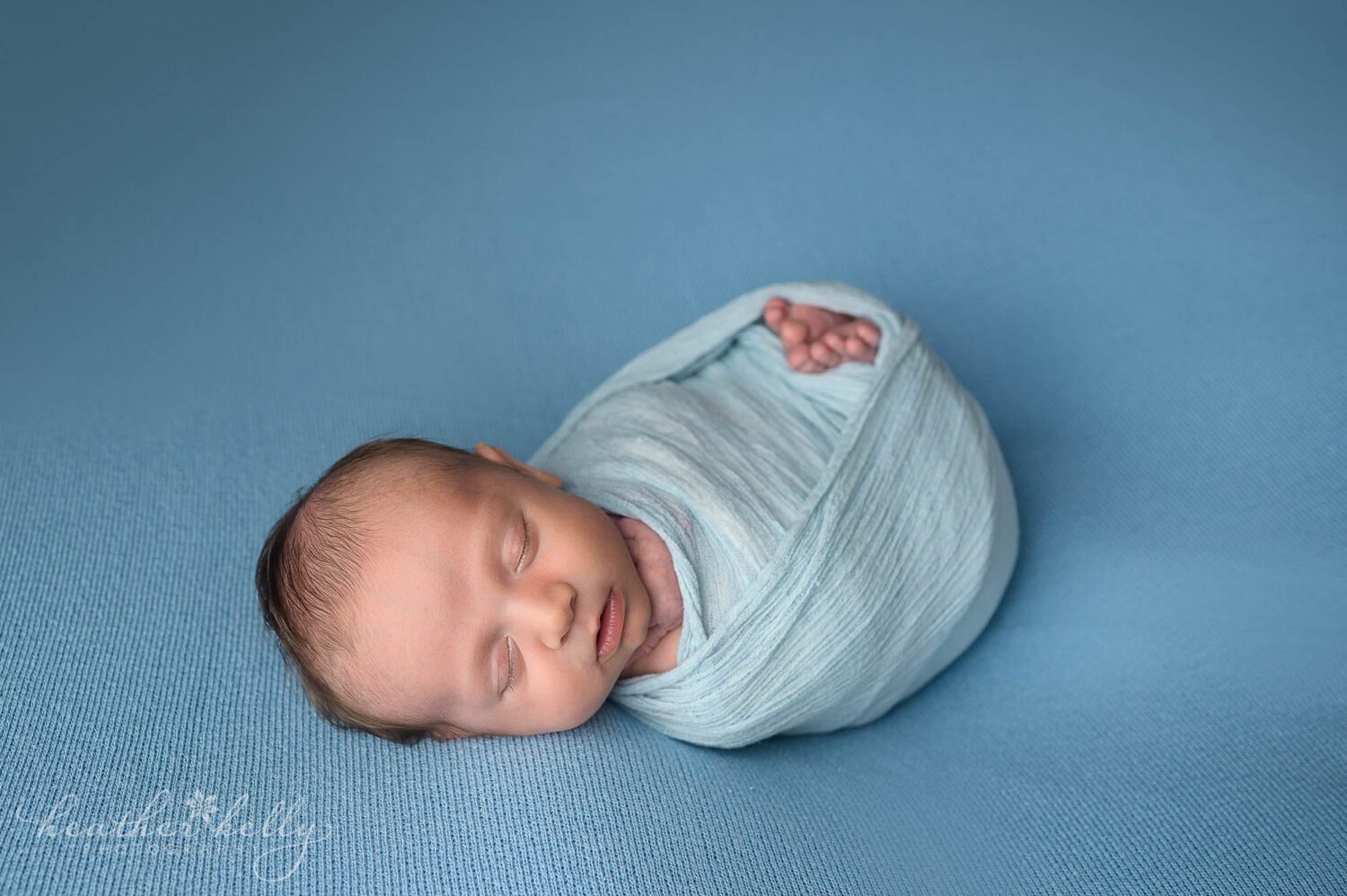 3 week old newborn boy posed for a newborn photography session in danbury ct. He is wrapped in blue with his toes peeking out and is laying on a blue backdrop. danbury ct newborn