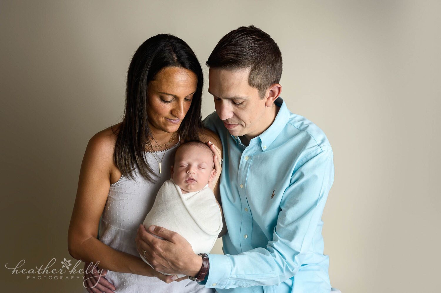 new mom and dad holding their newborn son. They are both looking down at him. He is wrapped in a white swaddle. newborn photography