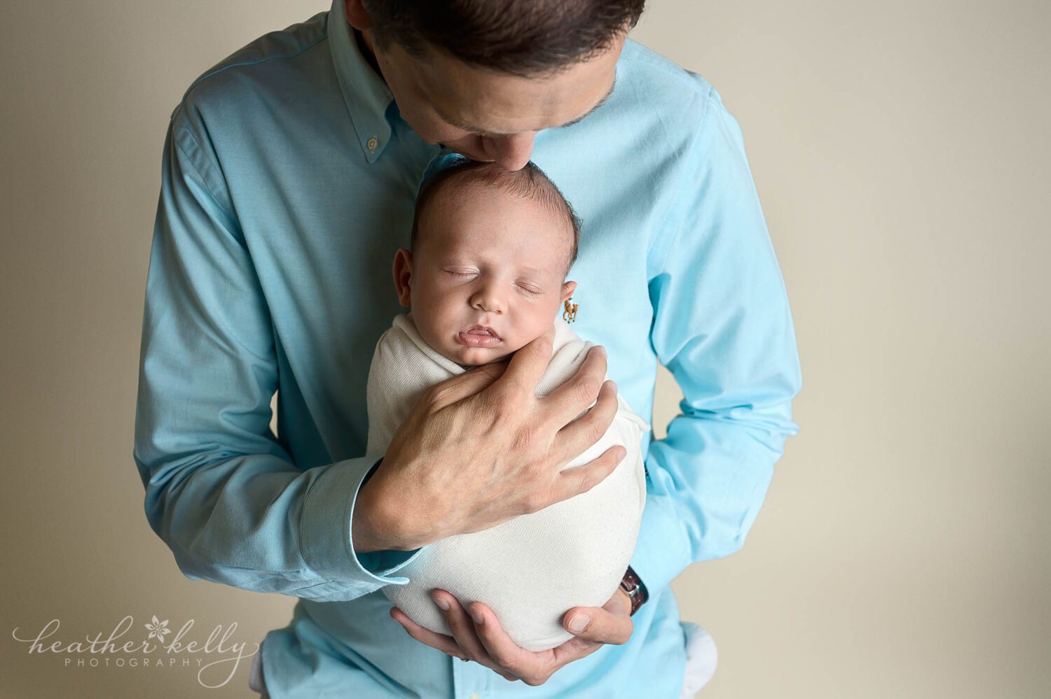 Dad is wearing a blue shirt and is holding his newborn son facing out against his chest. His head leans down and he is kissing his son and the forehead. 
