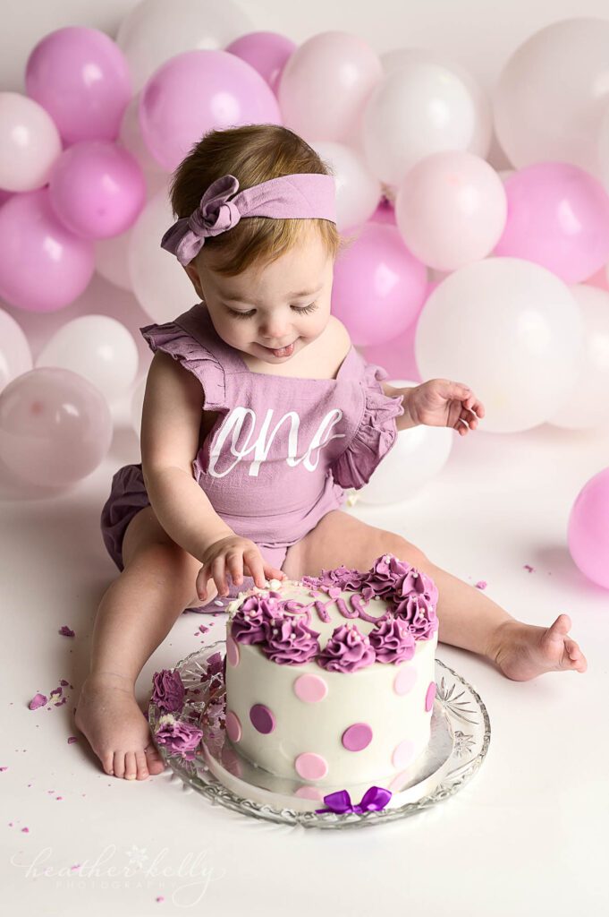 Ridgefield CT Cake Smash photography

A one year old girl in a purple outfit looking down and reaching out for her cake. There are different shades of white, pink, and purple balloons in the background. 