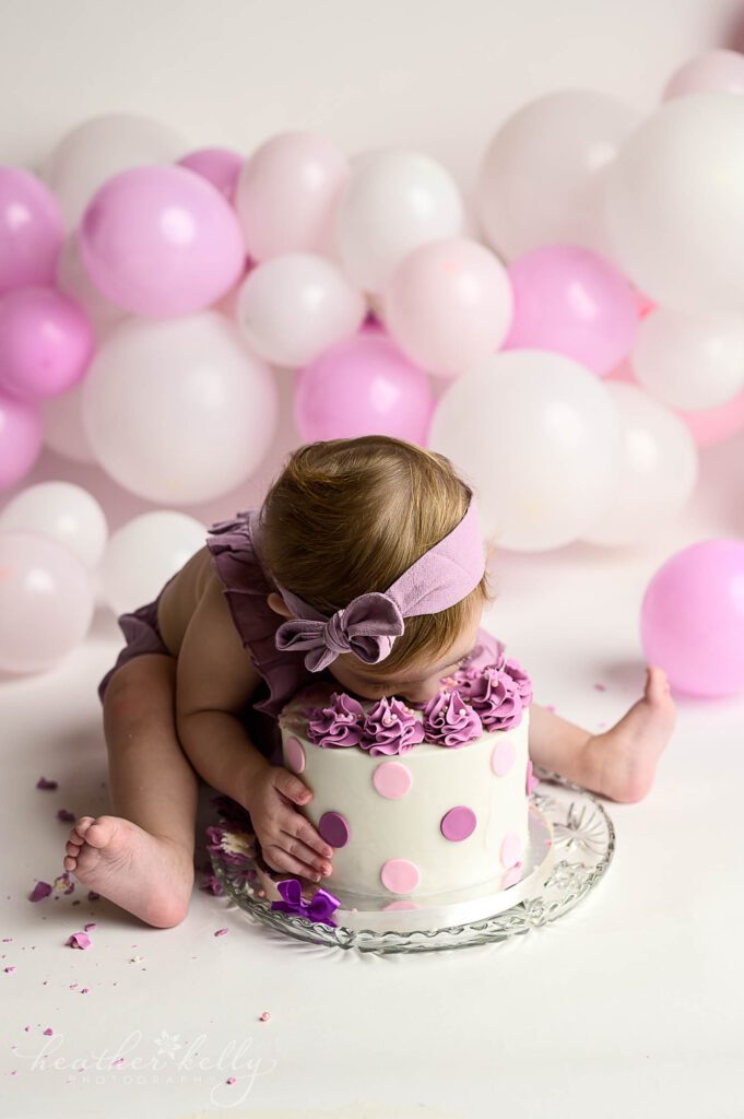 Ridgefield CT Cake Smash photography

A one year old girl in a purple outfit and headband, grabs hold of her cake on the floor and leans down to bite into it with her mouth. background of white, pink, and purple balloons. 