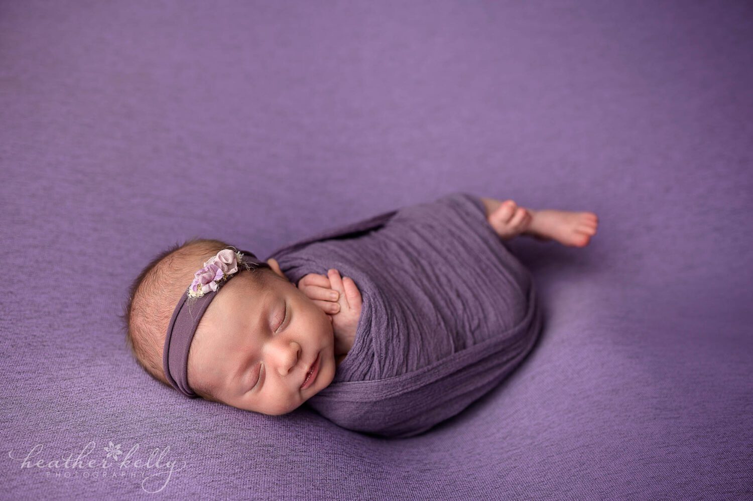 A newborn wrapped in purple with a purple headband. She is sleeping during her photography session. Her little fingers and toes pop out. 
