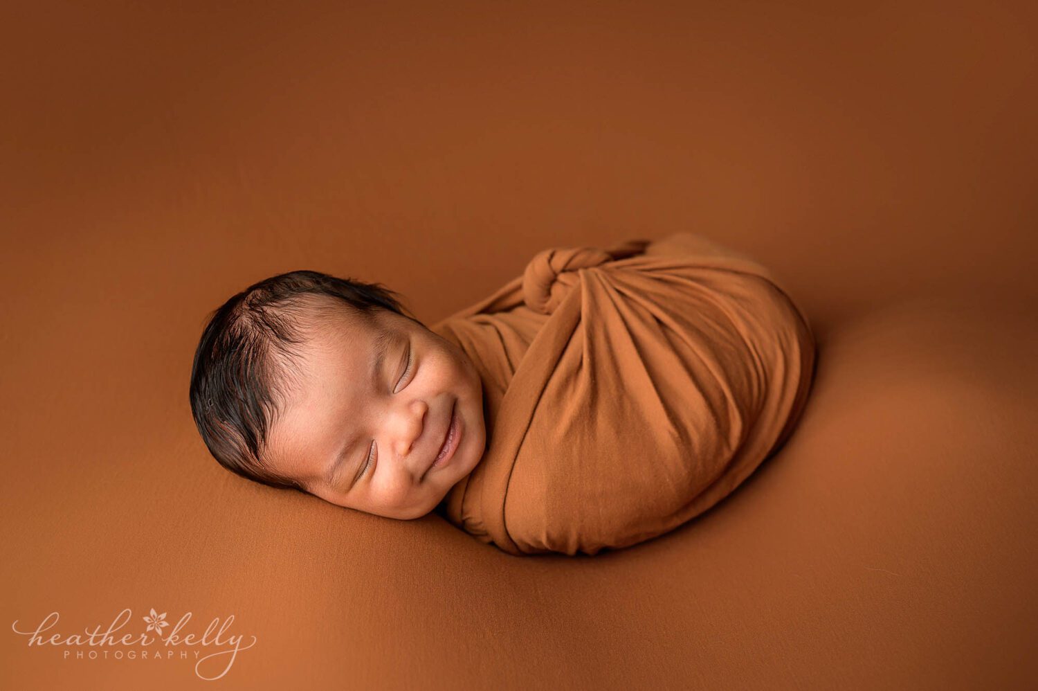 A newborn boy is wrapped in a burnt orange blanket and laying on an orange backdrop. He is sleeping and has a smile on his face. 