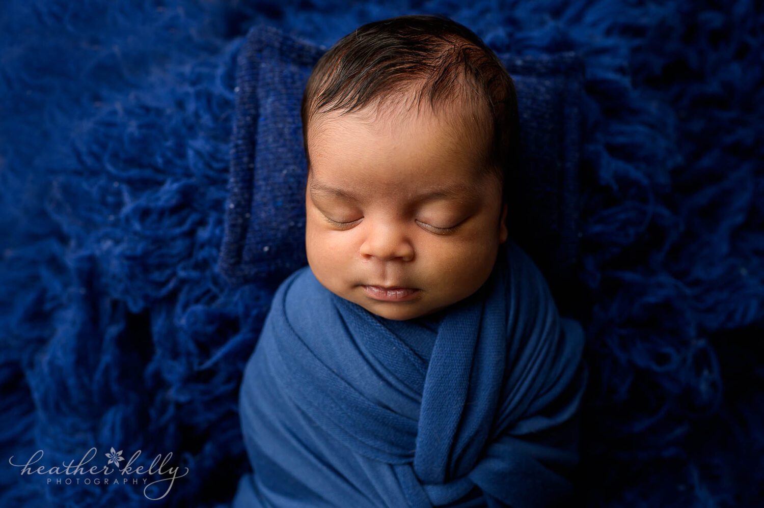 A baby boy is posed during a newborn photography session in newtown ct. He is wrapped in a navy blue swaddle and has a small blue pillow behind his head. He is on a blue flokati rug. 