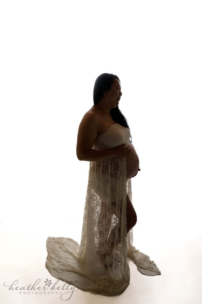 a mom to be wearing a white lace maternity gown during her maternity photography session. There is a high key white background. Danbury maternity photographer