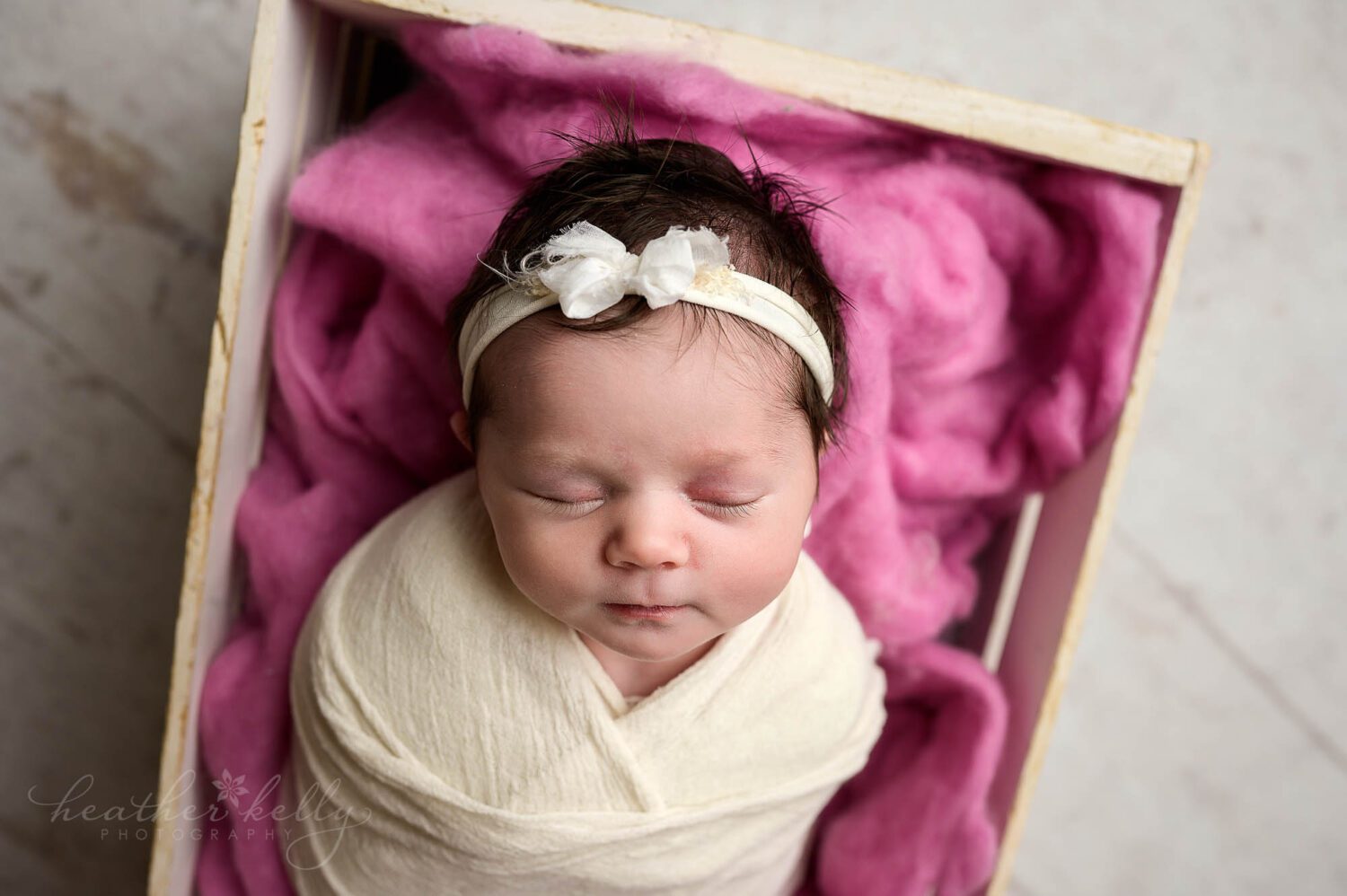 a newborn girl wrapped in cream. She is sleeping in a white crate with deep pink felt. 