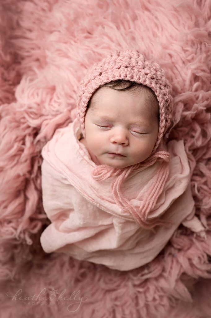 a newborn girl is wearing a pink bonnet. she is wrapped in pink and sleeping on a pink flokati rug 