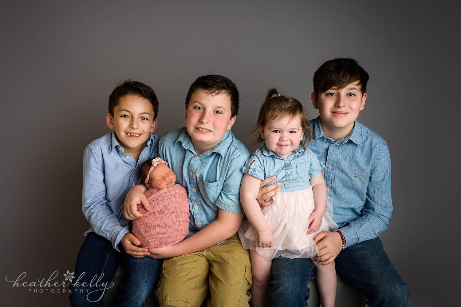 4 siblings with their newborn sister. They are sitting on a bench looking at the camera during their newborn photography session. 