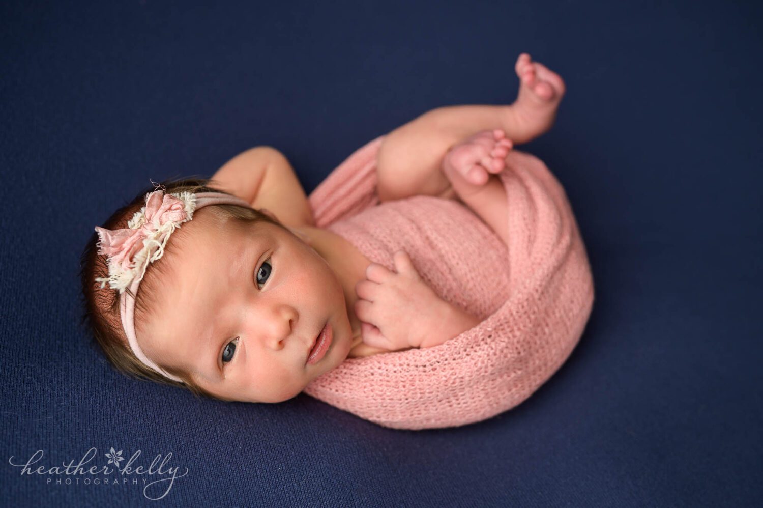 a newborn girl is partially wrapped in a pink wrap. Her hands and legs are sticking out. Her eyes are wide open while looking at the camera. She is laying on a blue backdrop. 