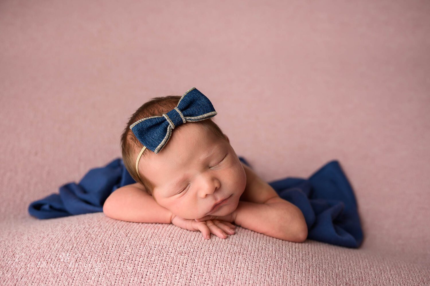 a chin up newborn pose of a ct newborn photography session. Baby girl is wearing a denim bow headband and on a pink backdrop. A blue wrap is draped over her. 