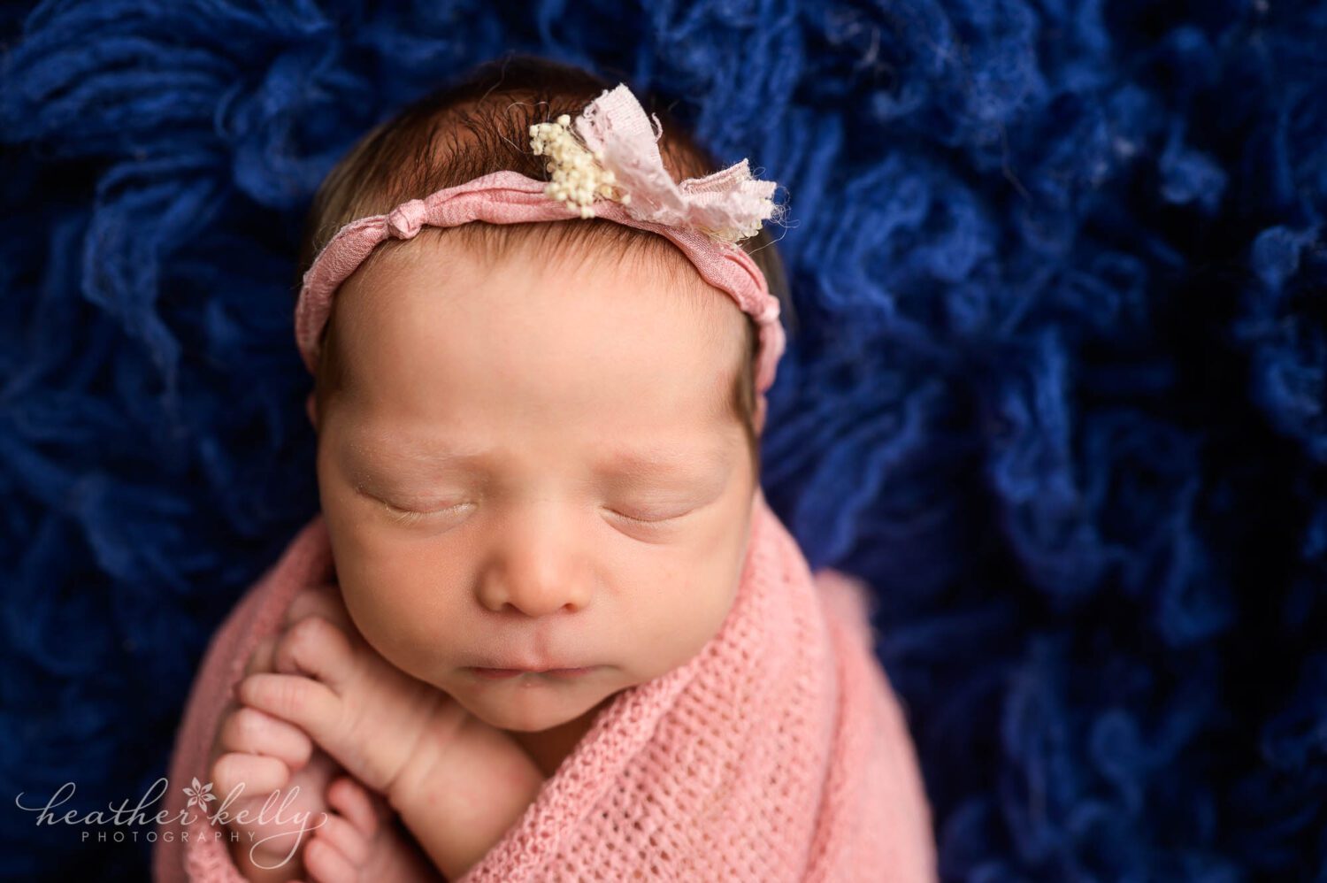 a newborn girl is wrapped in pink with her arms and toes sticking out. She is on a blue flokati rug. 