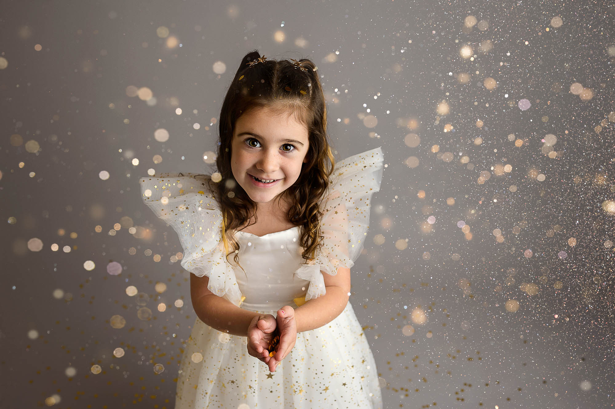 a 4 year old is wearing a white and gold sparkly dress. She is throwing gold glitter with a gray background.