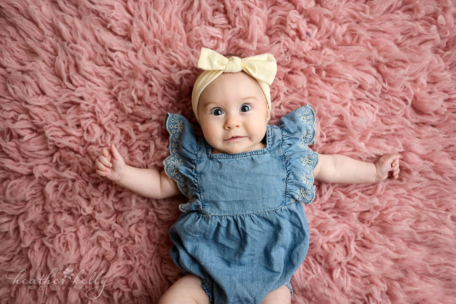 A baby girl is wearing a denim one piece outfit with ruffles on the short sleeves. She has a cream bow headband and is smiling at the camera. 