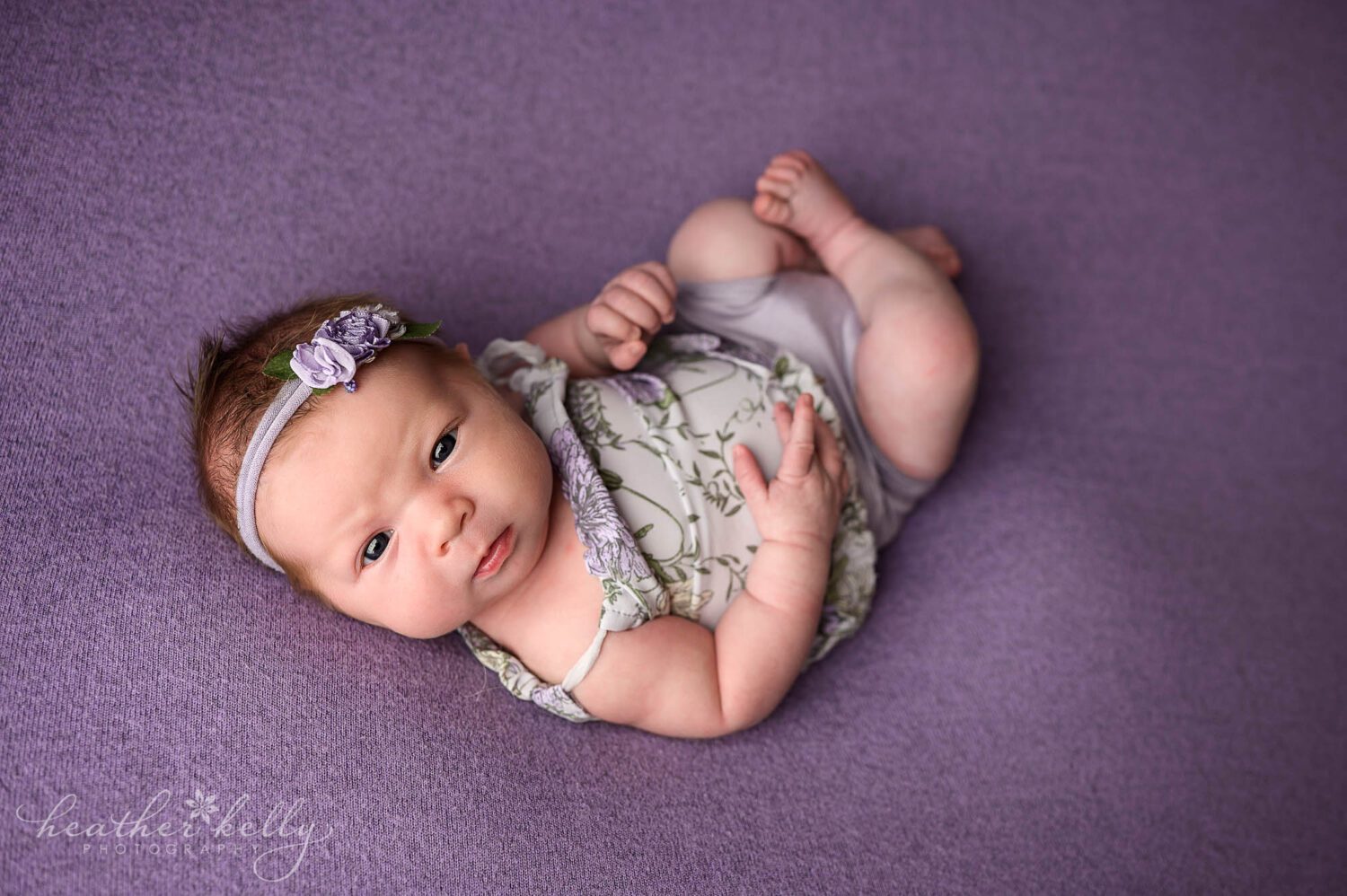 a newborn girl makes eye contact with the camera during her Newtown Ct newborn session. She is wearing a purple outfit and a purple flower headband. 